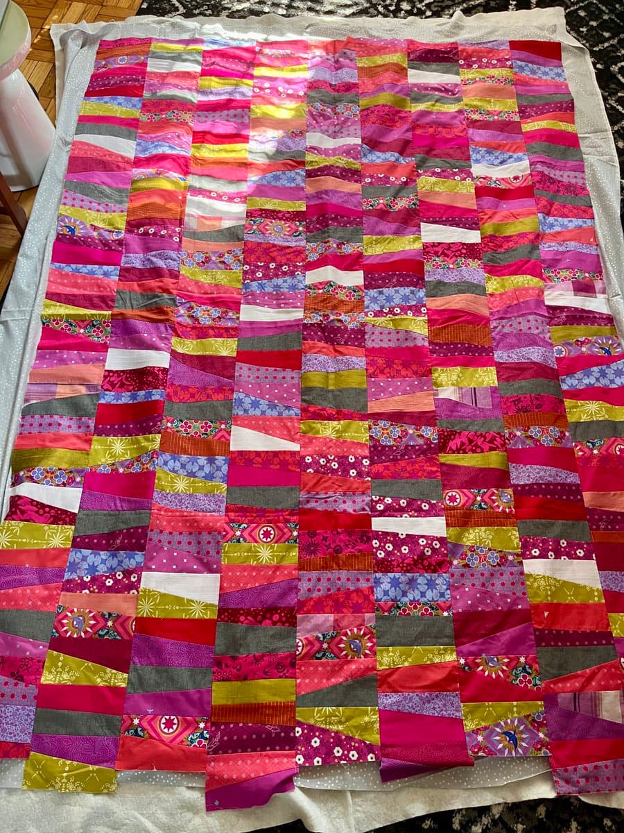 A colorful patchwork quilt made of horizontal fabric strips in shades of pink, red, purple, and yellow, laid flat on a white surface. The quilt features a variety of patterns and textures, including floral prints and solid colors, arranged in a grid-like design.