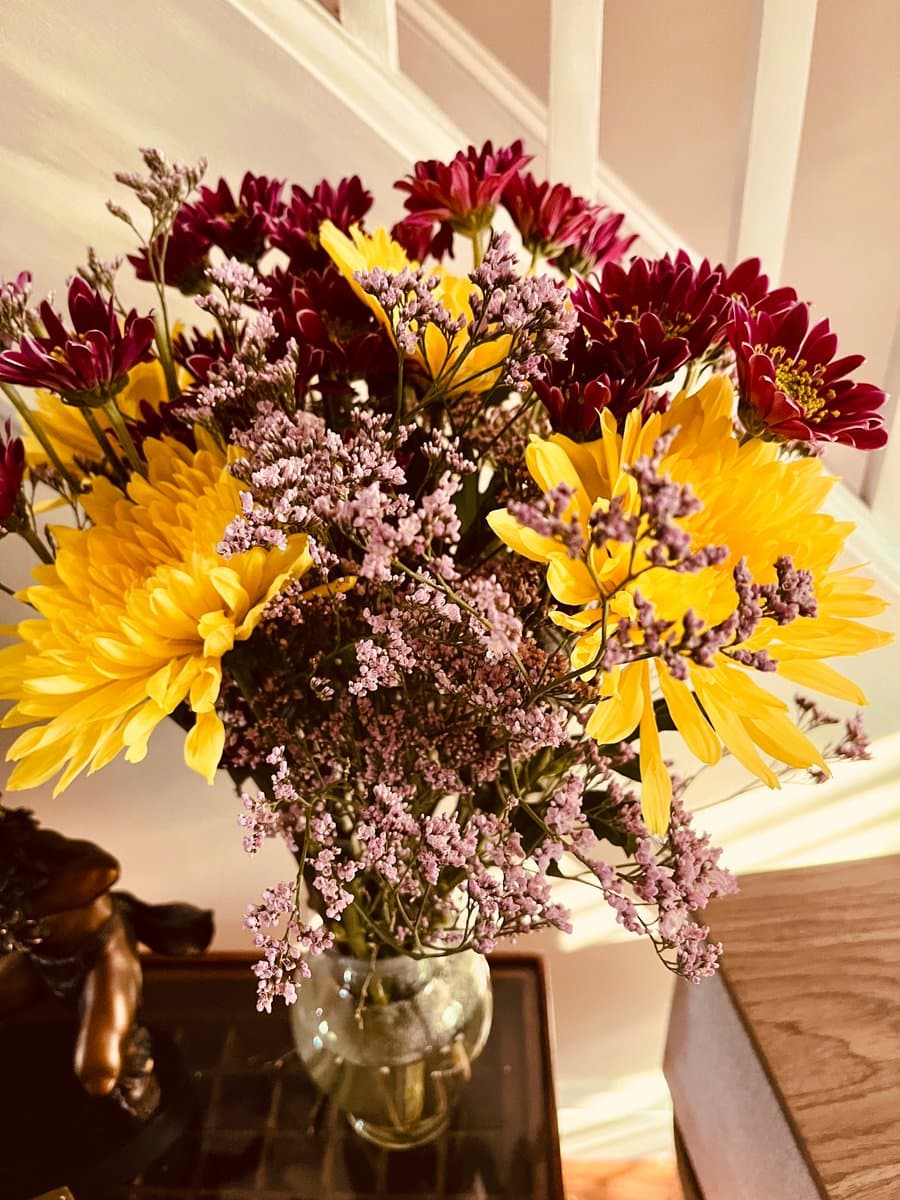 A close-up photograph of a vibrant flower arrangement in a clear glass vase, featuring large yellow and deep red chrysanthemums mixed with sprigs of small pink flowers. The vase sits on a dark, reflective surface next to a wooden piece of furniture, with a white wall and door frame in the background.