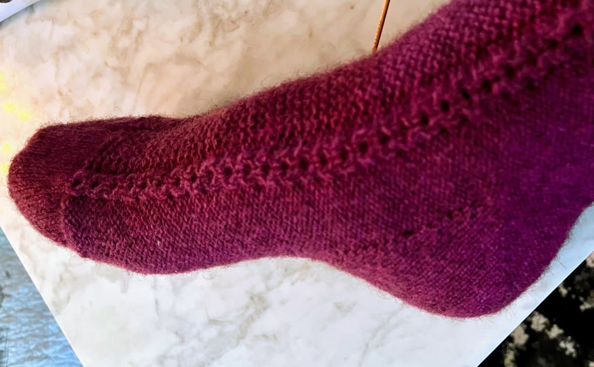 A close-up photograph of a hand-knitted sock in a deep magenta color, featuring a textured stitch pattern, resting on a white surface.