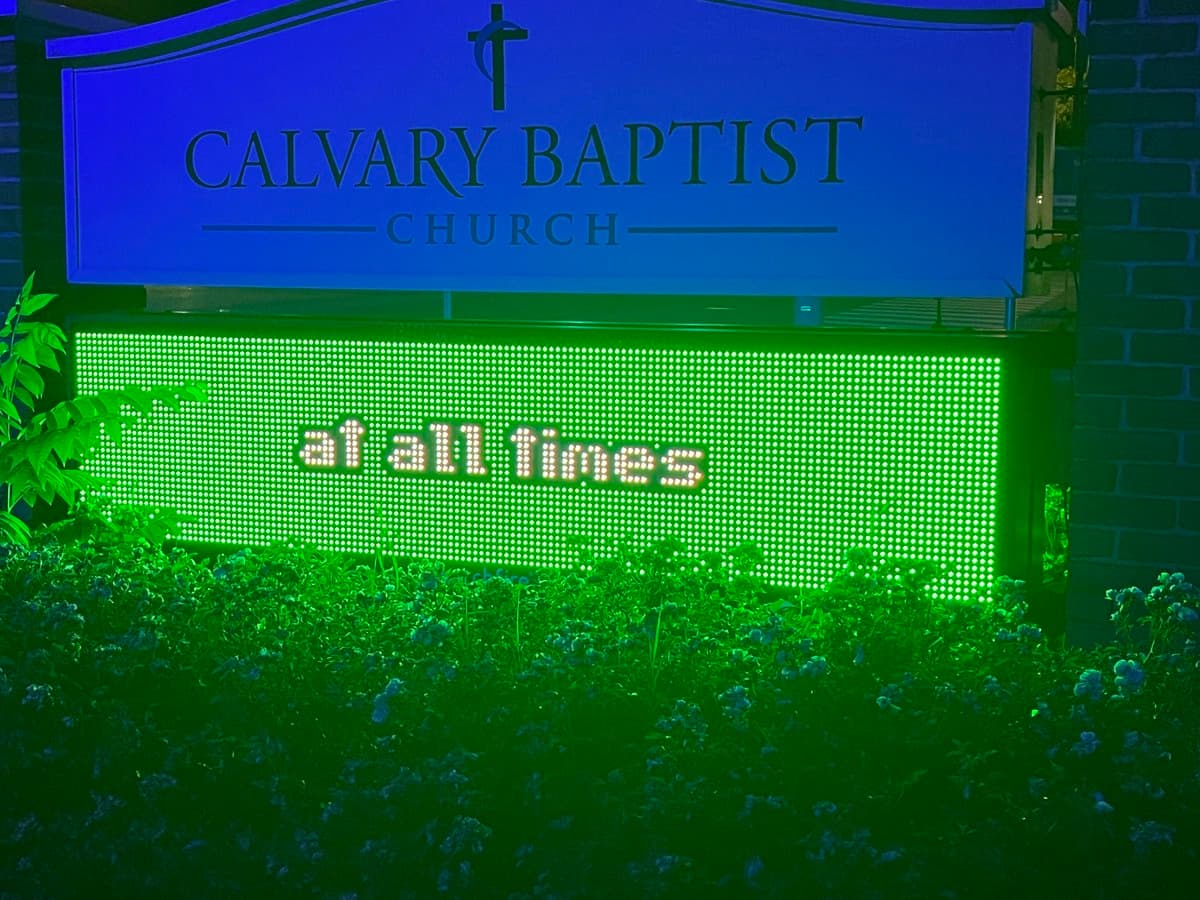 A nighttime photo of a church sign for Calvary Baptist Church, featuring a blue upper panel with the church name and a green LED display below it. The LED screen shows the text 'at all times' in white letters, illuminated against the dark background and surrounding greenery.