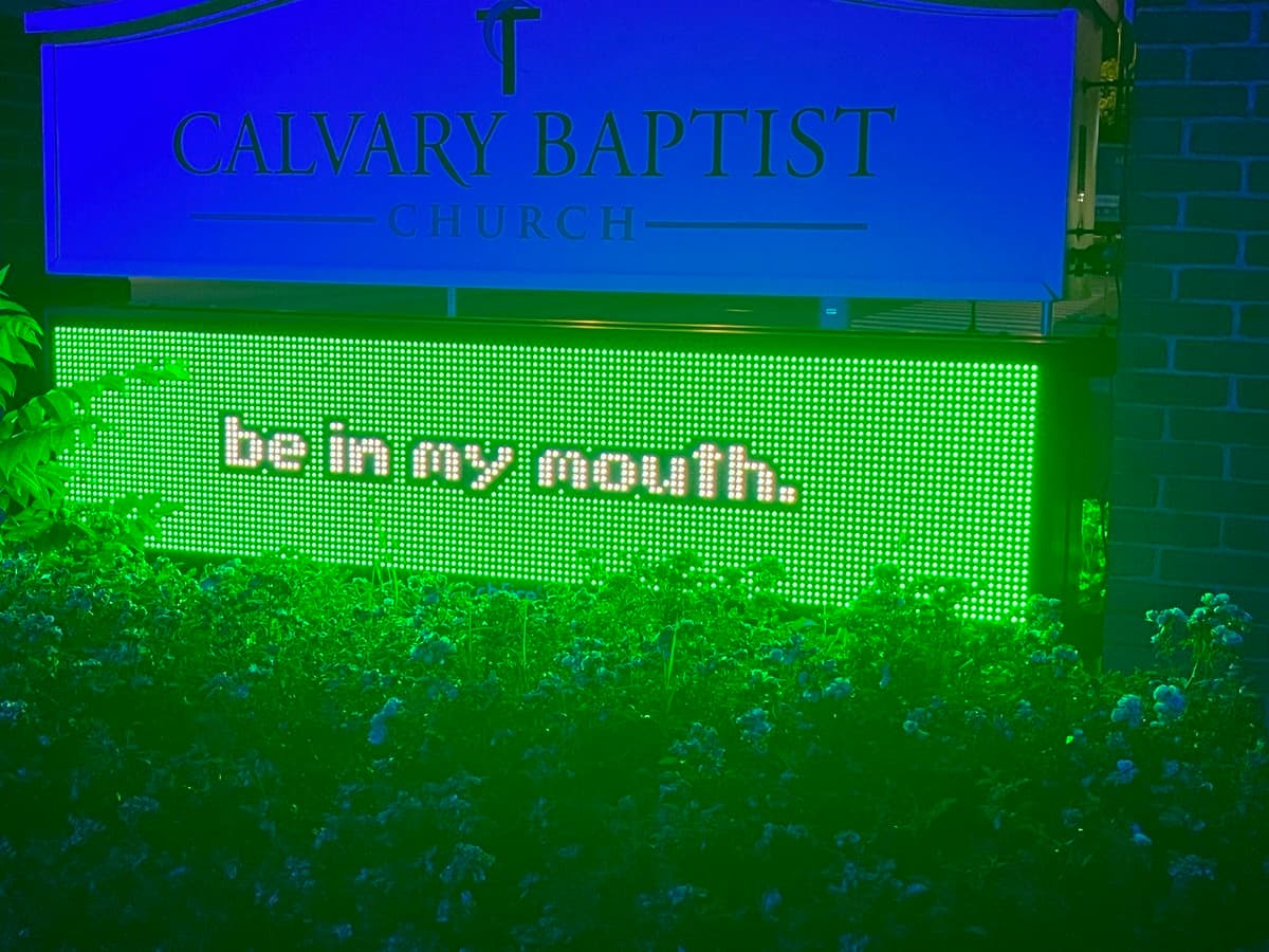 A nighttime photo of a church sign for Calvary Baptist Church, featuring a blue header and a green LED display below it. The LED screen shows the text 'be in my mouth.' in white letters, surrounded by illuminated bushes.
