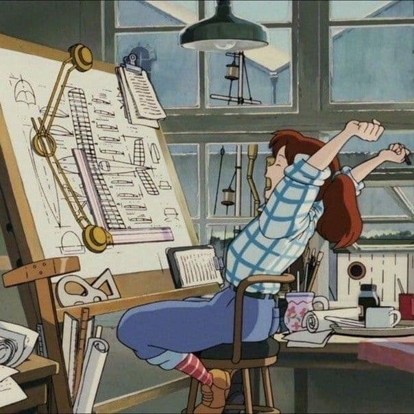A still from an animated film showing a young woman with red hair in a blue plaid shirt stretching her arms while sitting at a cluttered artist's desk. The desk holds an easel with technical drawings, a lamp, and various art supplies, set against a backdrop of large windows.