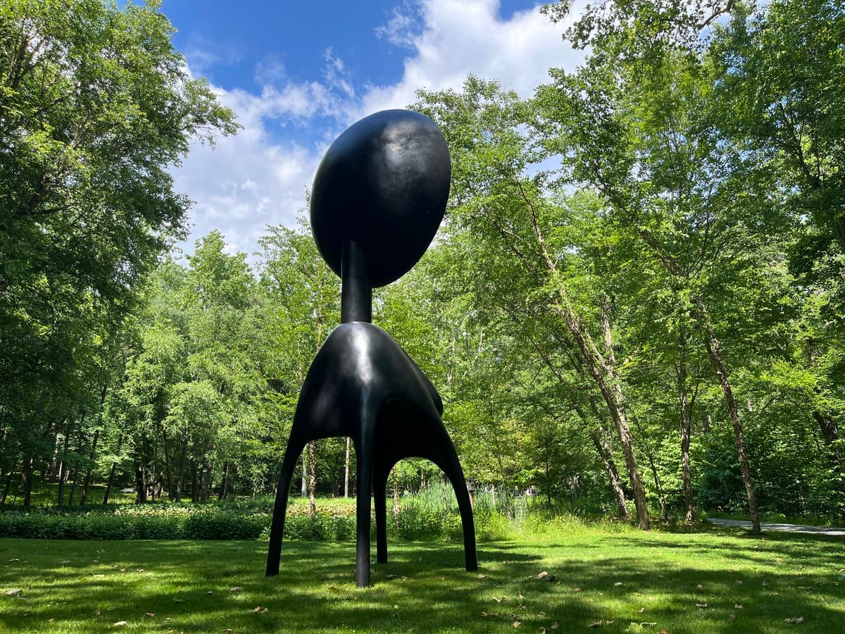 A black, abstract outdoor sculpture with a large spherical head and a slender, multi-legged body stands on a grassy lawn surrounded by trees under a blue sky with white clouds.