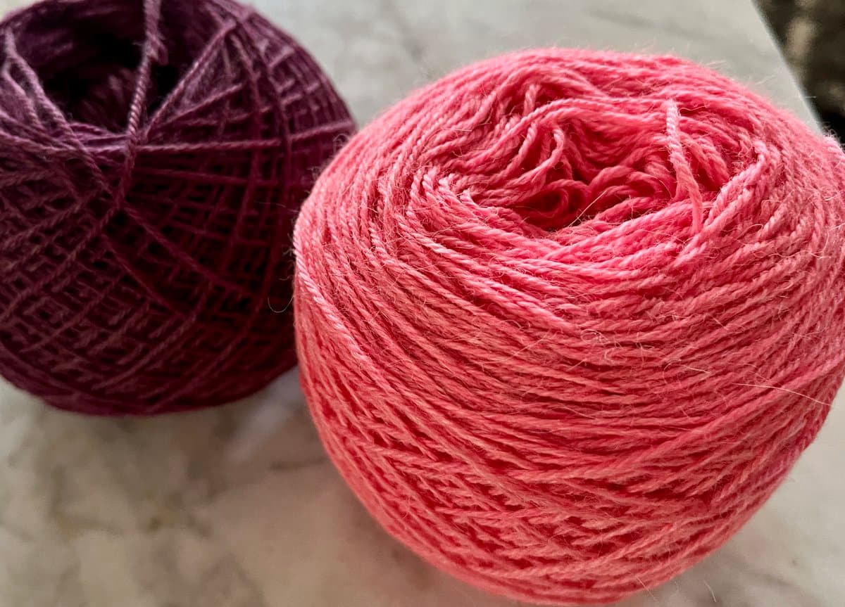 A close-up photograph of two balls of yarn resting on a light-colored surface. The ball in the foreground is a bright pink, while the ball in the background is a deep purple.