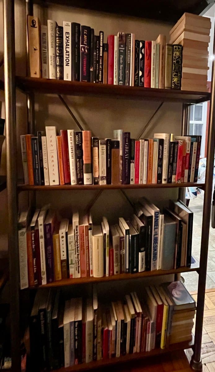 A photograph of a four-tiered wooden bookshelf filled with numerous books of various sizes and colors. The shelves are packed with literature, and a stack of books is placed horizontally on the top right corner.