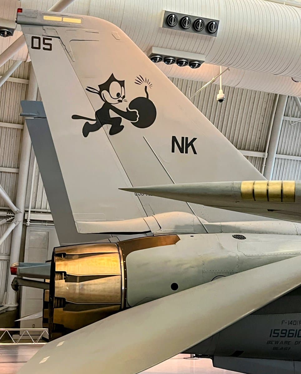 A close-up photograph of the tail section of a military aircraft displayed indoors, featuring a painted cartoon cat mascot holding a bomb, the letters "NK", and the number "05" on the vertical stabilizer.