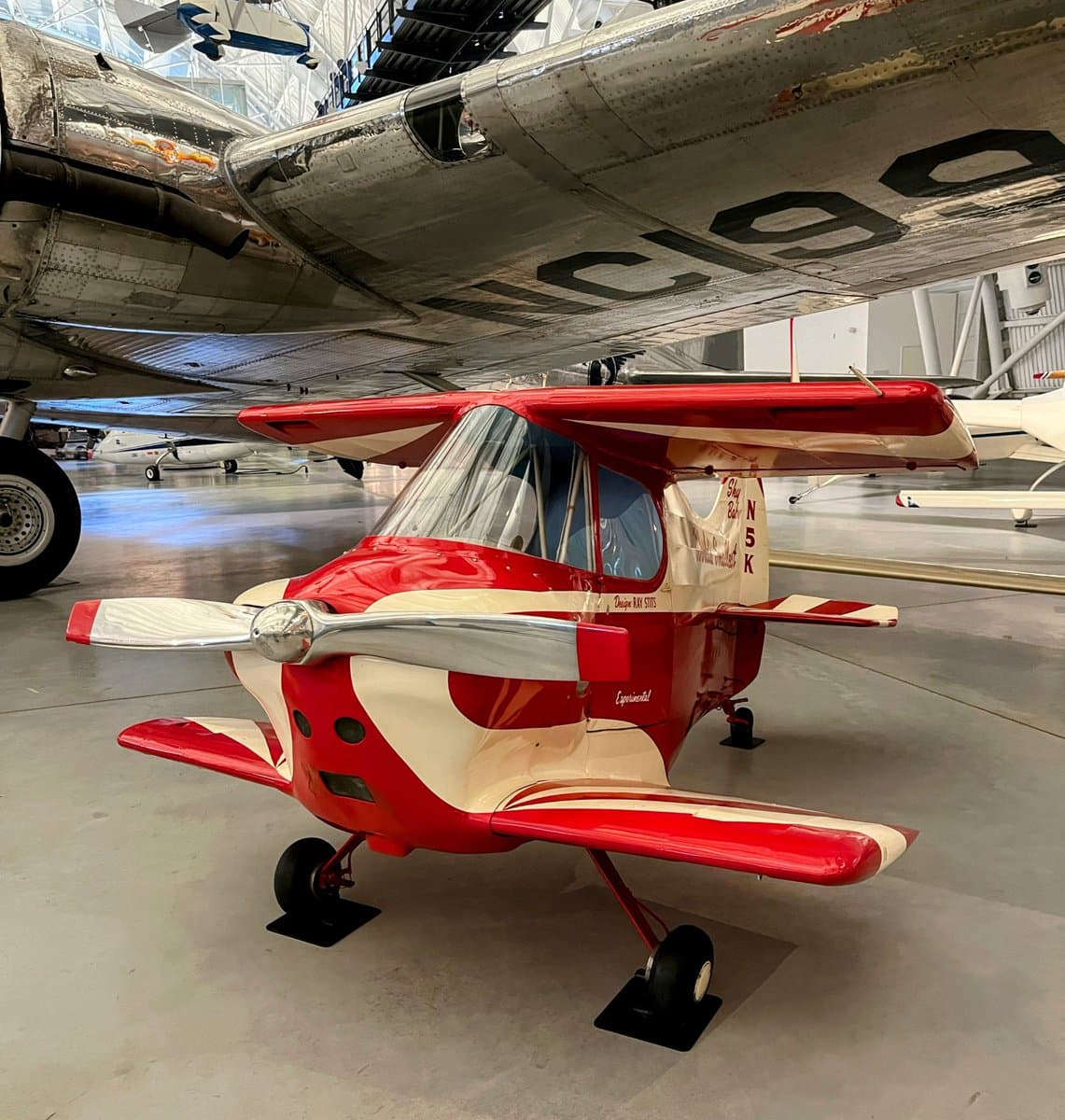 A red and white experimental aircraft is displayed on the floor of a museum hangar, positioned beneath the wing of a large silver airplane.