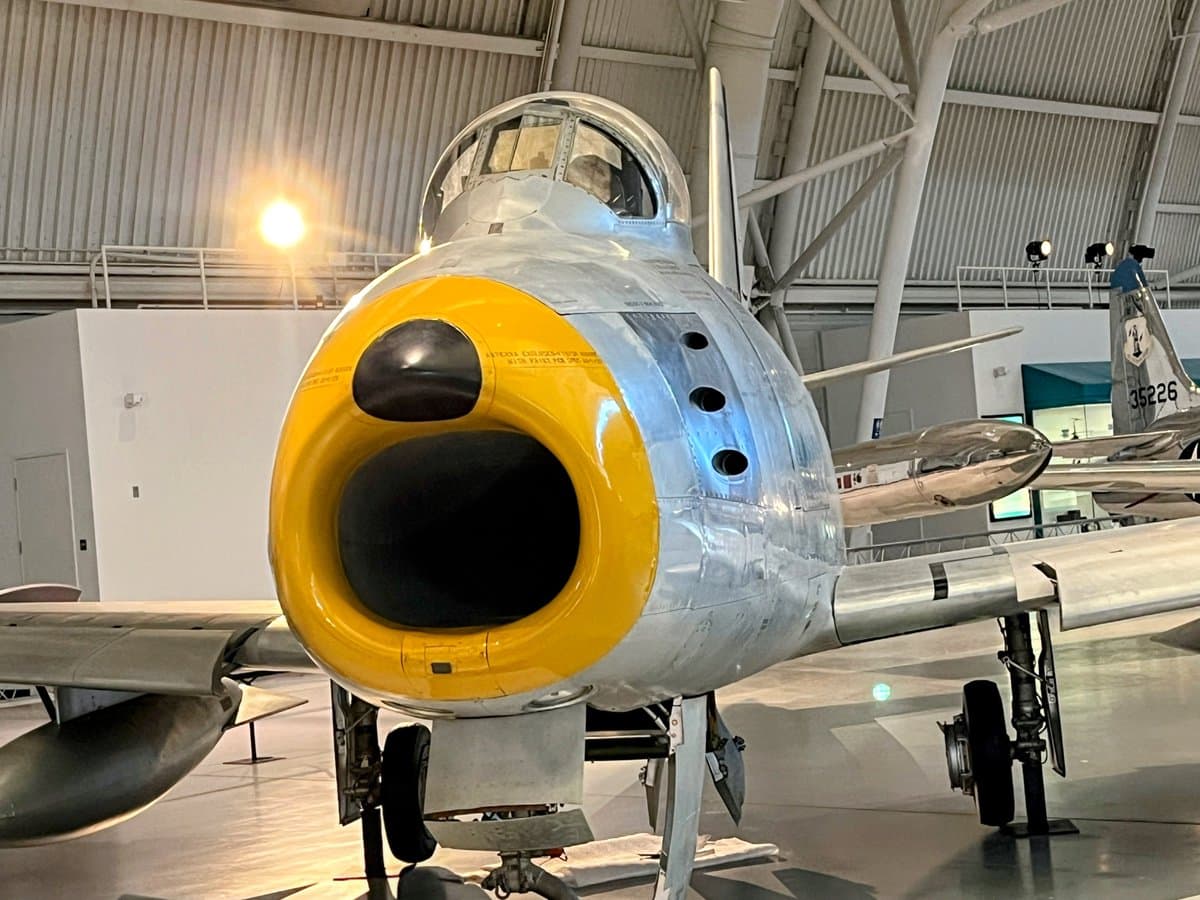A front-facing view of a silver military jet aircraft with a distinctive yellow nose intake, displayed indoors in a museum hangar. The aircraft features a bubble canopy and is parked on its landing gear, with another aircraft visible in the background.
