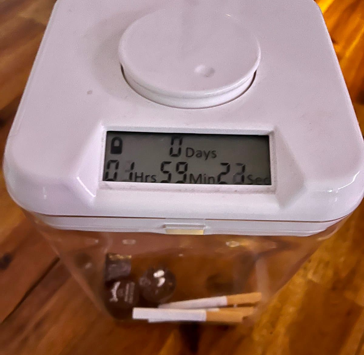 A close-up photograph of a white digital timer sitting on a wooden surface, with a clear plastic container underneath holding three peanut butter cups and two cigarettes. The timer's display shows a countdown of 0 days, 1 hour, 59 minutes, and 22 seconds.
