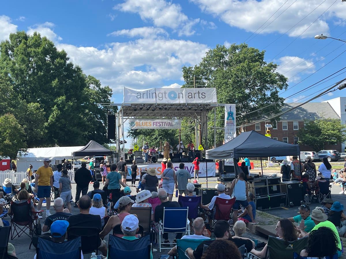 An outdoor daytime photograph of a crowd seated in folding chairs watching a band perform on a stage at the Columbia Pike Blues Festival. The stage is covered by a banner reading "arlington arts" and "BLUES FESTIVAL," with trees and a blue sky in the background.