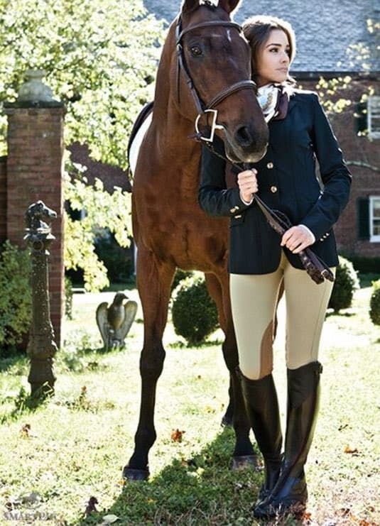 A woman in equestrian attire stands beside a brown horse in a grassy outdoor setting. She is wearing a black riding jacket, beige breeches, and tall black riding boots, holding the horse's reins. The background features a brick building, trees, and garden decorations.