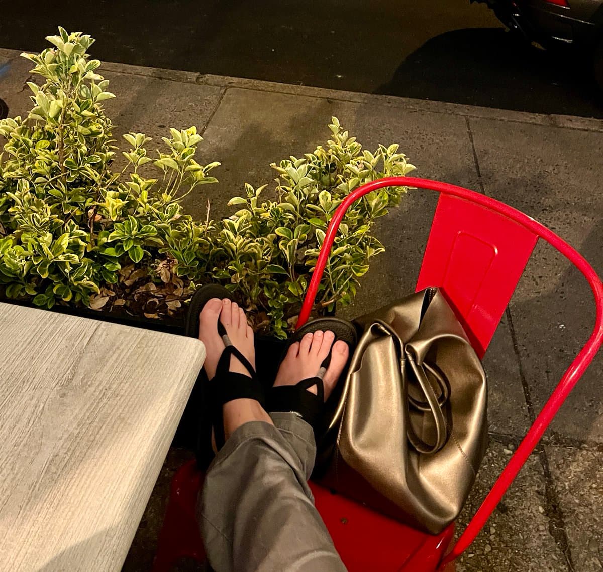 A first-person view of a person's feet in black flip-flops resting on a red metal chair, with a metallic gold handbag placed beside them. The scene is set outdoors at night on a paved surface next to a low green hedge and a light-colored table.