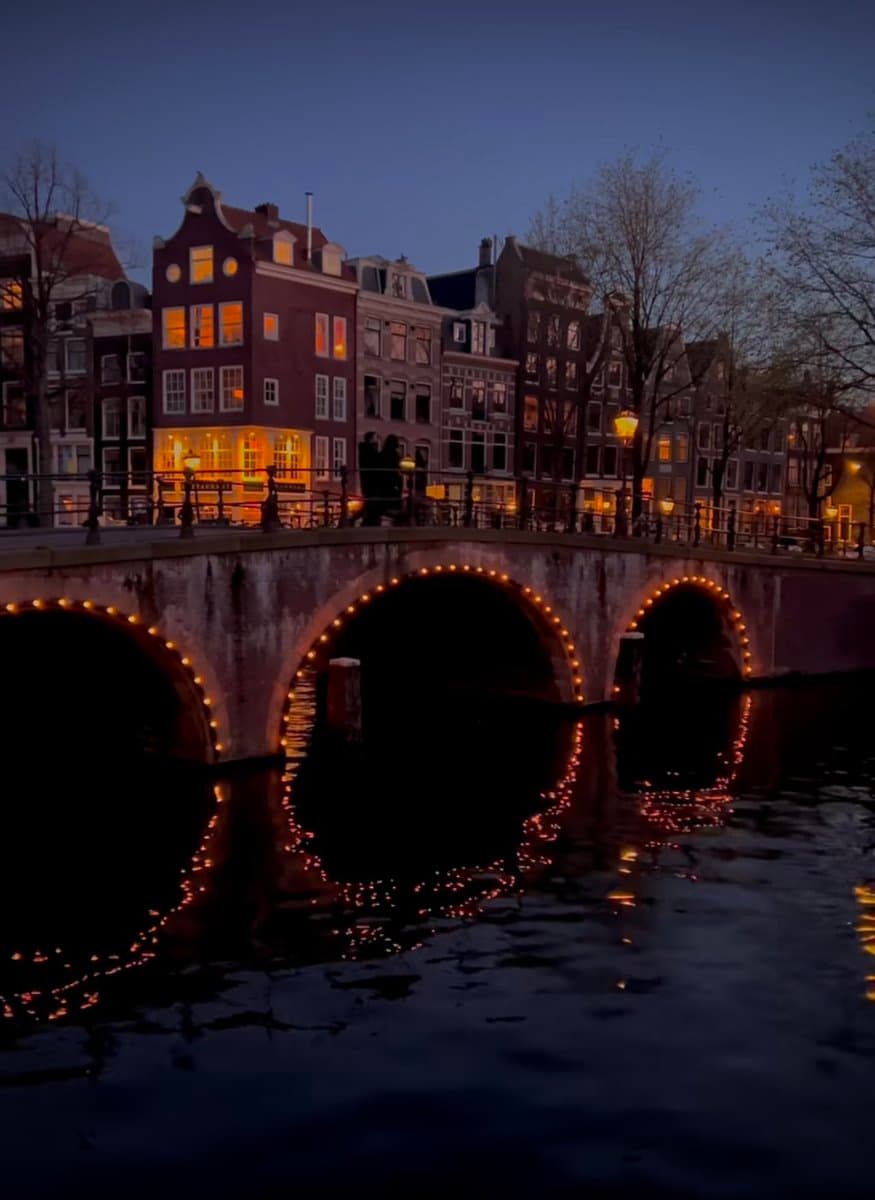 A night photograph of a canal in a European city, likely Amsterdam, featuring a stone bridge with three arches outlined in string lights. The lights reflect on the dark water below, while traditional canal houses with illuminated windows line the background.
