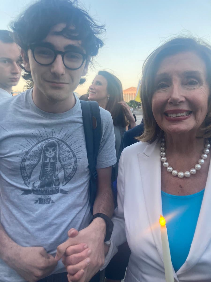 A selfie-style photograph of a young man with glasses and a grey t-shirt standing next to an older woman in a white blazer, who is holding a lit candle. They are outdoors with other people and a classical building visible in the background.