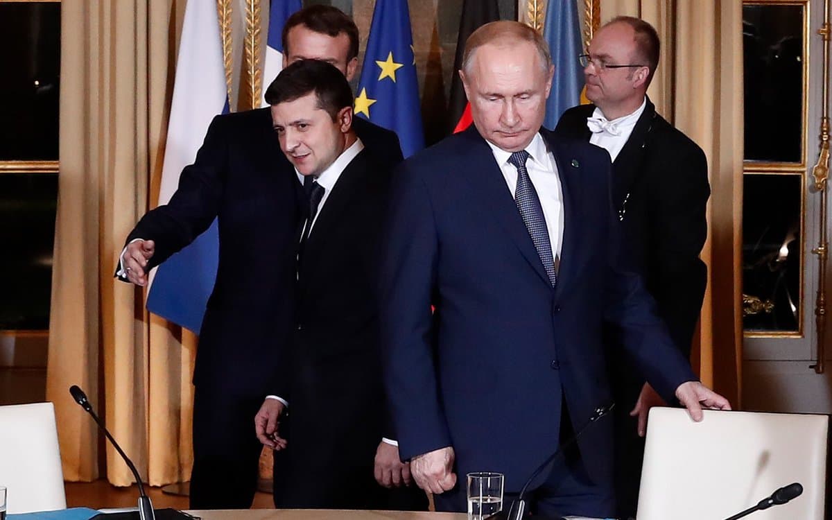 A photograph of four men in suits standing behind a table with microphones, with flags of France, the European Union, and Russia visible in the background.