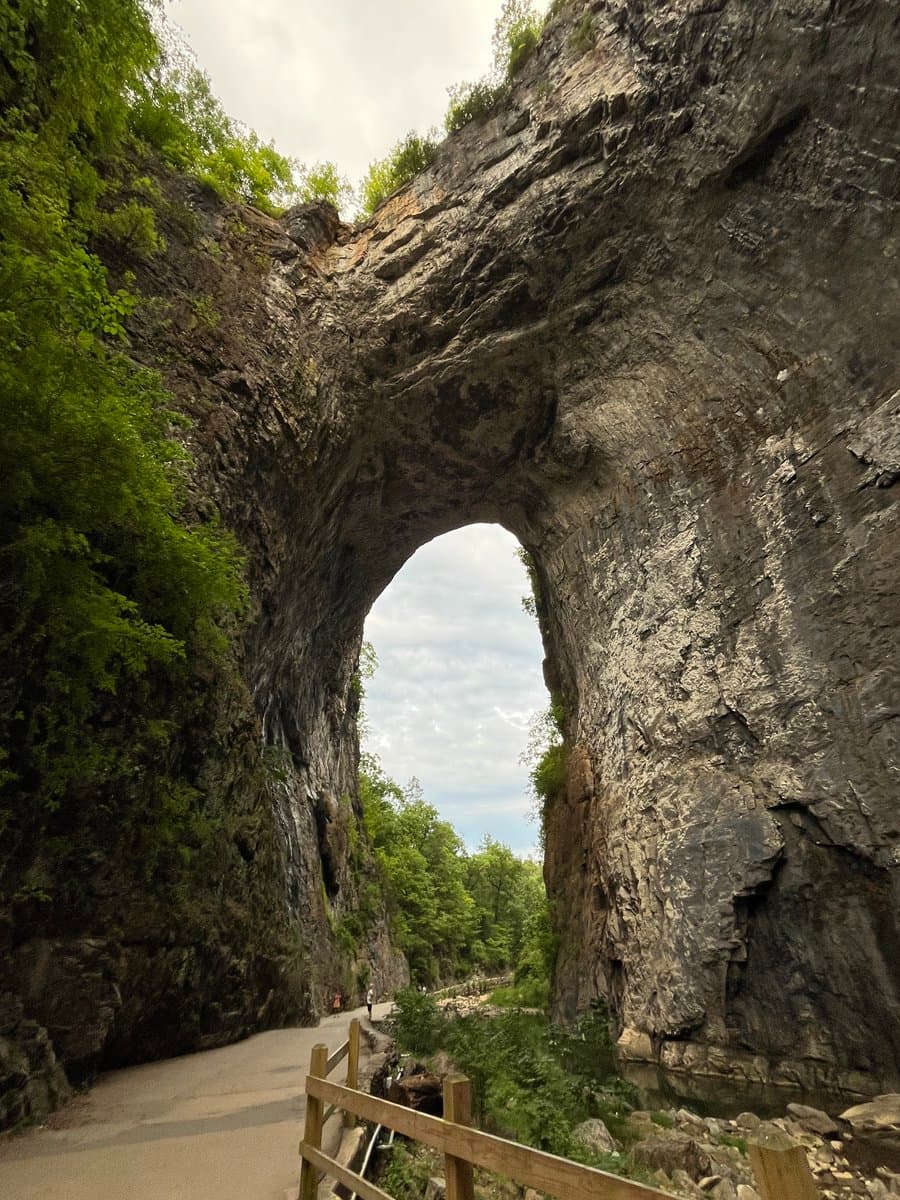A low-angle outdoor photograph of a large, natural stone archway covered in patches of green vegetation, with a paved walking path and wooden railing in the foreground.