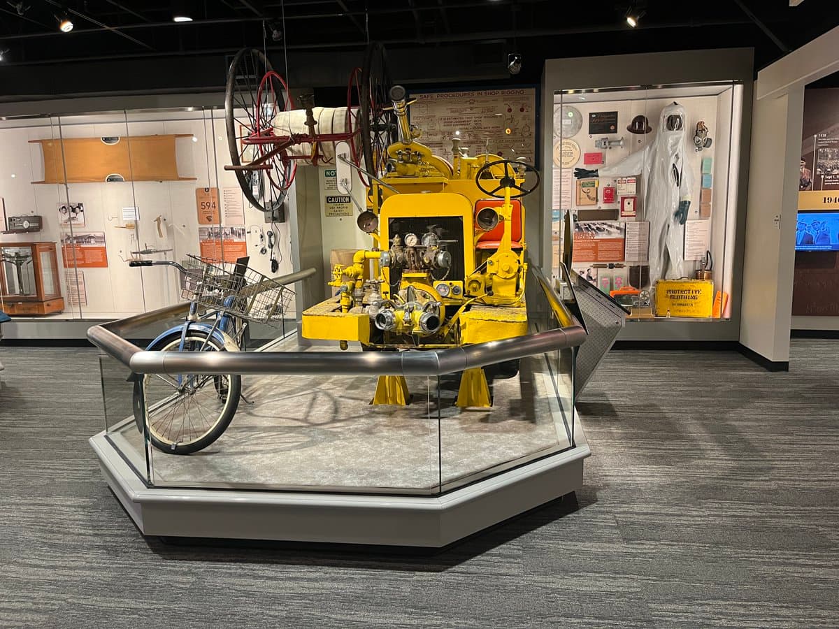 A museum exhibit featuring a large, yellow industrial machine with gauges and a steering wheel, displayed on a glass platform next to a blue bicycle. The background includes informational panels and a white protective suit in a display case.