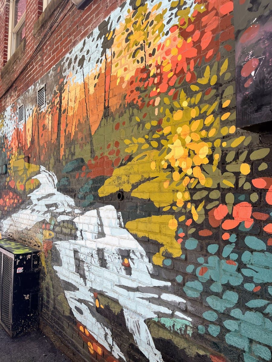 A colorful mural painted on a brick wall, depicting a landscape with autumn foliage in shades of yellow, orange, and red, along with a white, textured foreground. The artwork is located in an alleyway, with a black utility box visible on the left side.