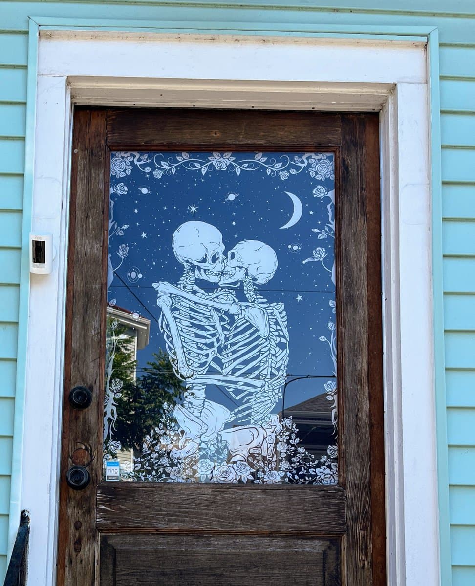 A photograph of a wooden front door featuring a large, white etched glass panel depicting two skeletons kissing under a starry night sky with a crescent moon. The door is set into a light blue house exterior, and a Ring doorbell is visible on the left side of the frame.