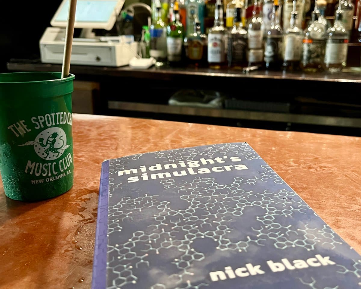 A copy of the novel "Midnight's Simulacra" by Nick Black sits on a bar counter next to a green drink cup from The Spotted Cat Music Club in New Orleans. In the background, a row of liquor bottles lines the back bar.