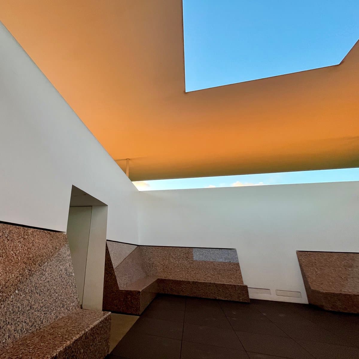 A low-angle architectural photograph of a modern, minimalist courtyard or rooftop space. The scene features white walls, a textured stone bench, and a large opening in the ceiling that reveals a blue sky, with warm light illuminating the interior surfaces.