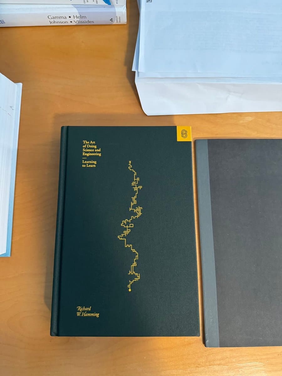 A photograph of a black hardcover book titled "The Art of Doing Science and Engineering: Learning to Learn" by Richard W. Hamming, resting on a wooden desk next to a stack of papers and a gray notebook. The book cover features a gold, pixelated line graph and a yellow tab with the letter 'S'.