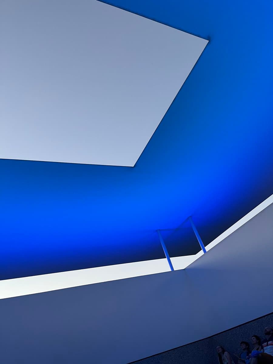 A low-angle architectural photograph looking up at a modern building's interior, featuring a bright white skylight and a deep blue ceiling illuminated by artificial light. A small group of people is visible sitting on a ledge in the bottom right corner.