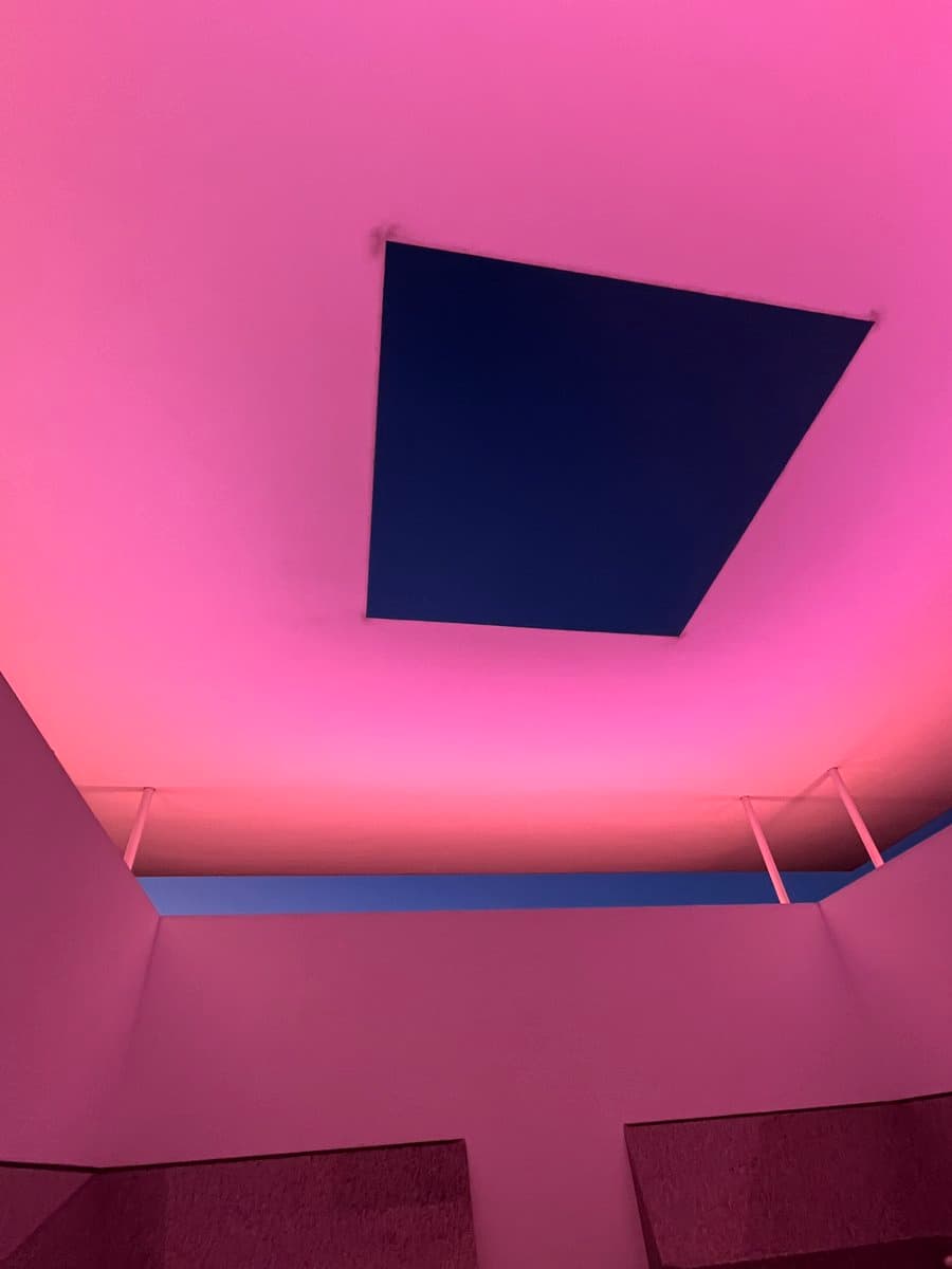 A low-angle photograph looking up at a modern architectural structure, featuring a large, dark square skylight set into a ceiling that is illuminated with a vibrant pink and purple light. Below the ceiling, white walls and thin support poles frame a strip of blue sky, creating a minimalist and geometric composition.