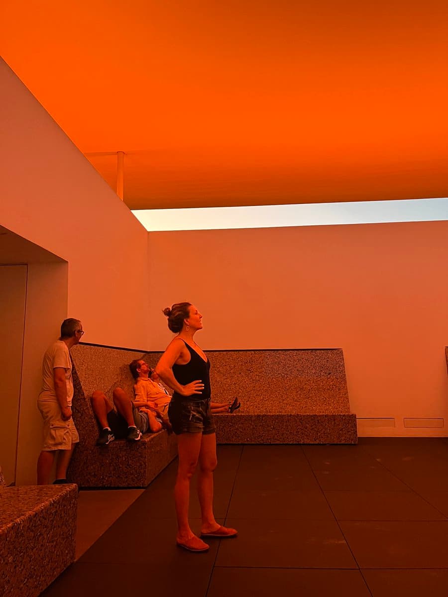 A photograph of a modern, minimalist interior space bathed in a warm, orange light, likely from a skylight or colored lighting. A woman stands in the foreground looking up, while two other people rest on a long stone bench in the background.