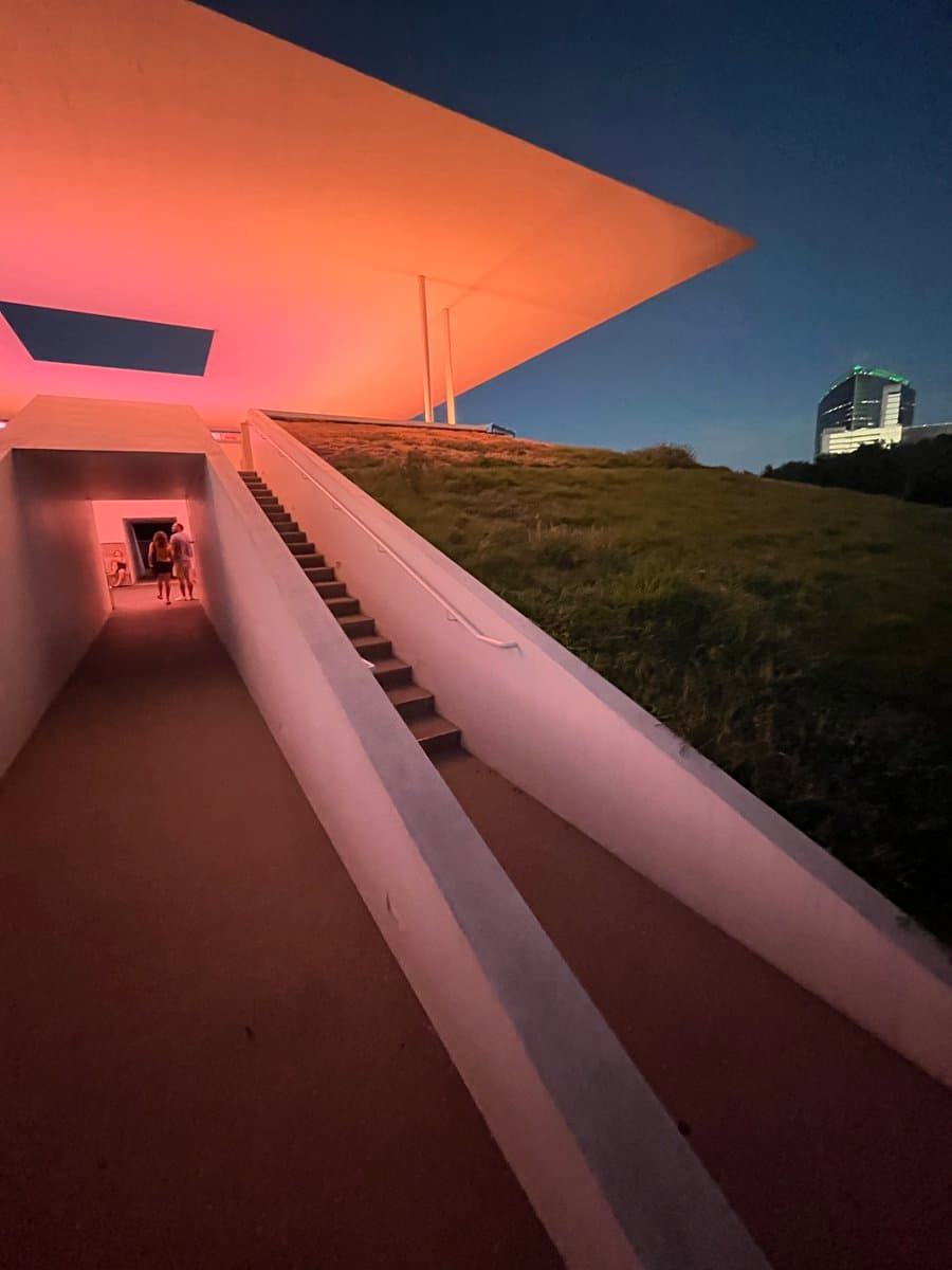 A low-angle photograph of a modern architectural structure at dusk, featuring a large, flat roof illuminated with pink and orange light. A concrete staircase and ramp lead up to the building, with a grassy hill and a city skyline visible in the background under a dark blue sky.