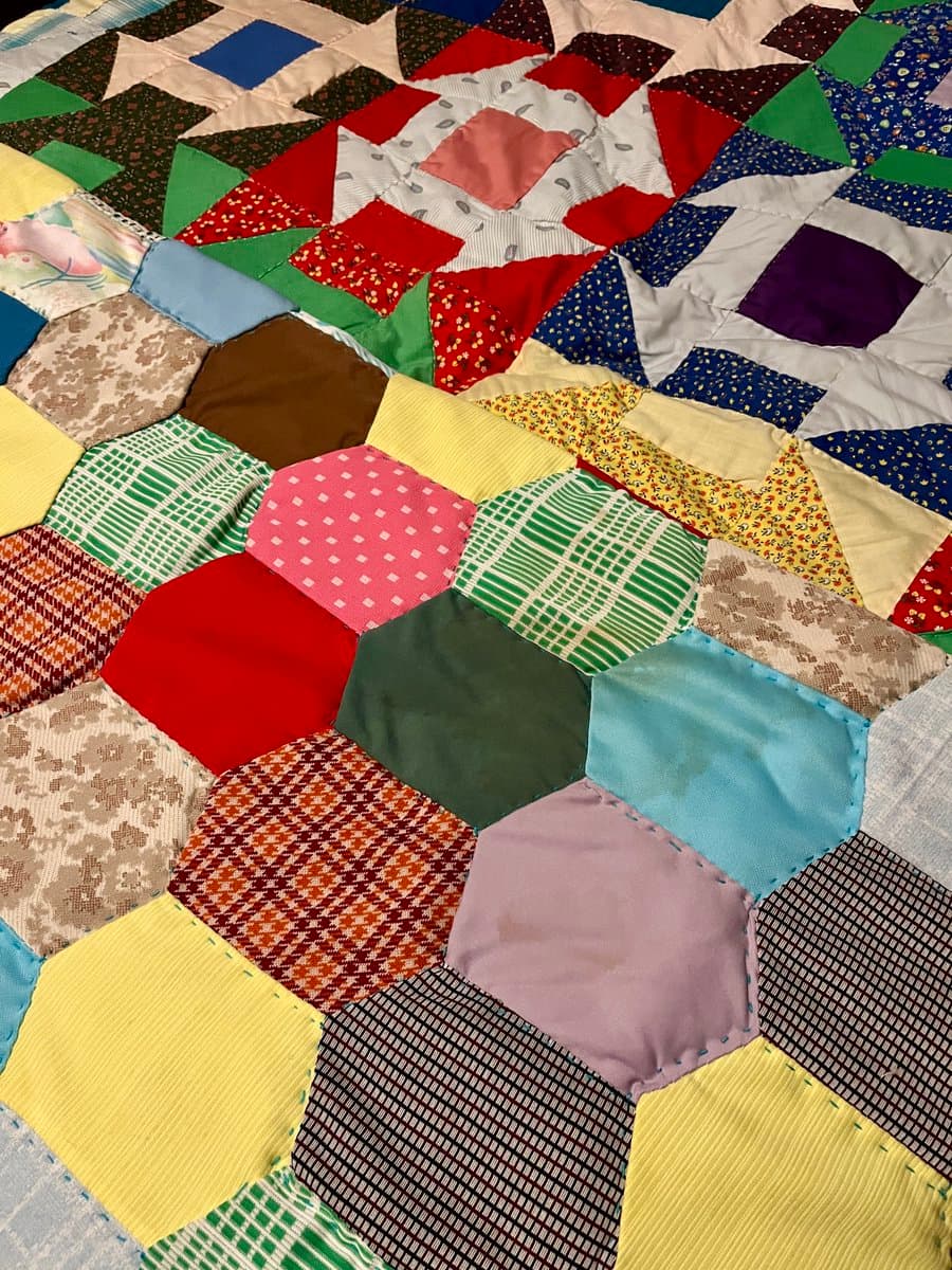 A close-up view of a colorful, hand-sewn patchwork quilt featuring a variety of geometric shapes and patterns. The fabric pieces include hexagons, triangles, and squares in diverse colors and prints, stitched together with visible seams.