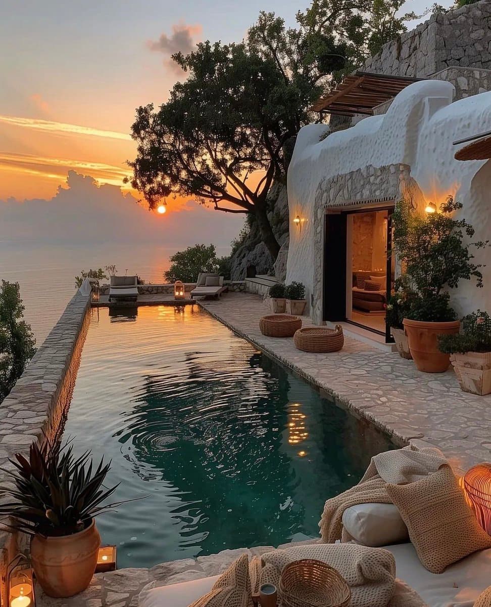 A scenic photograph of a luxury cliffside villa featuring a long, narrow infinity pool overlooking the ocean at sunset. The white-washed building has an open doorway revealing a warmly lit interior, and the poolside is decorated with potted plants, woven baskets, and lounge chairs.