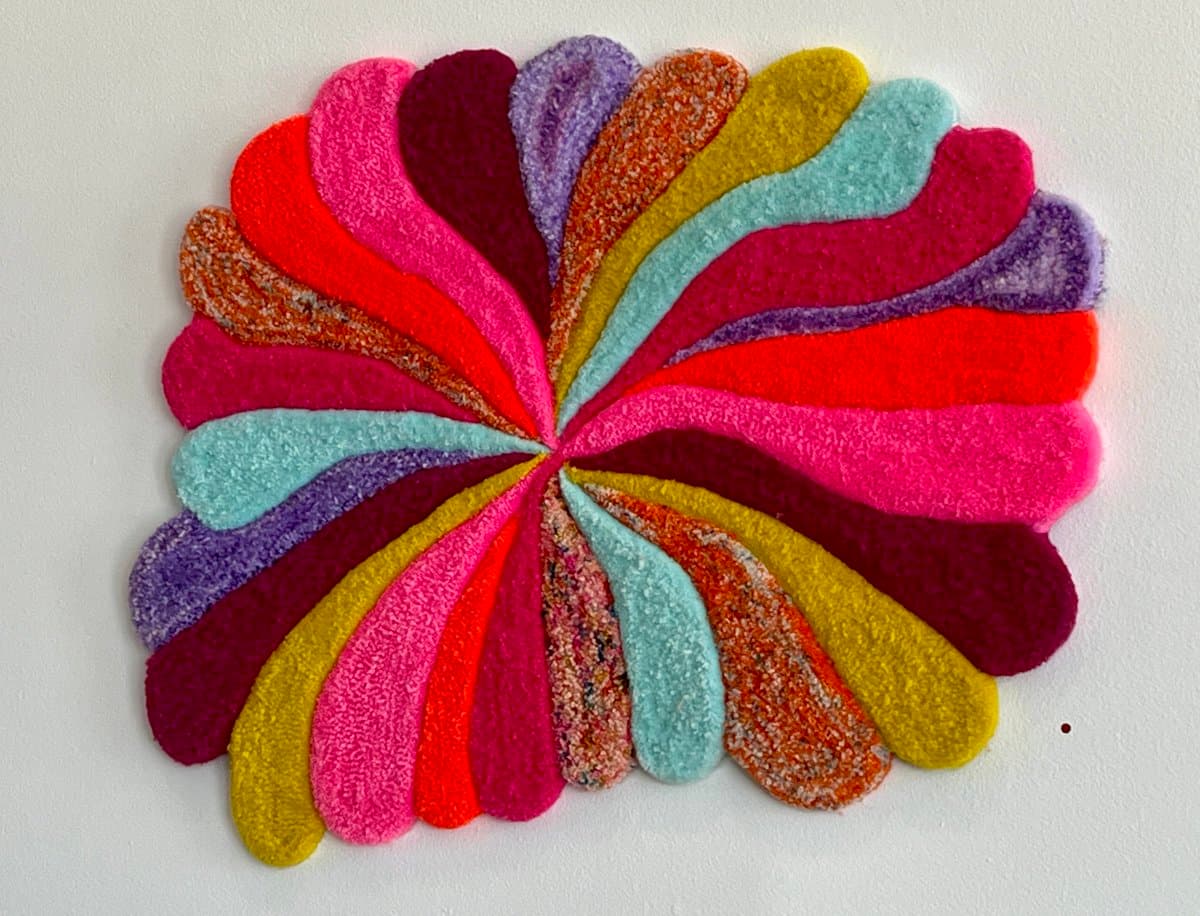 A colorful, circular felt craft made of multicolored, petal-shaped pieces arranged in a pinwheel pattern on a white background. The pieces are in shades of pink, red, orange, yellow, purple, and blue, with some featuring a speckled texture.