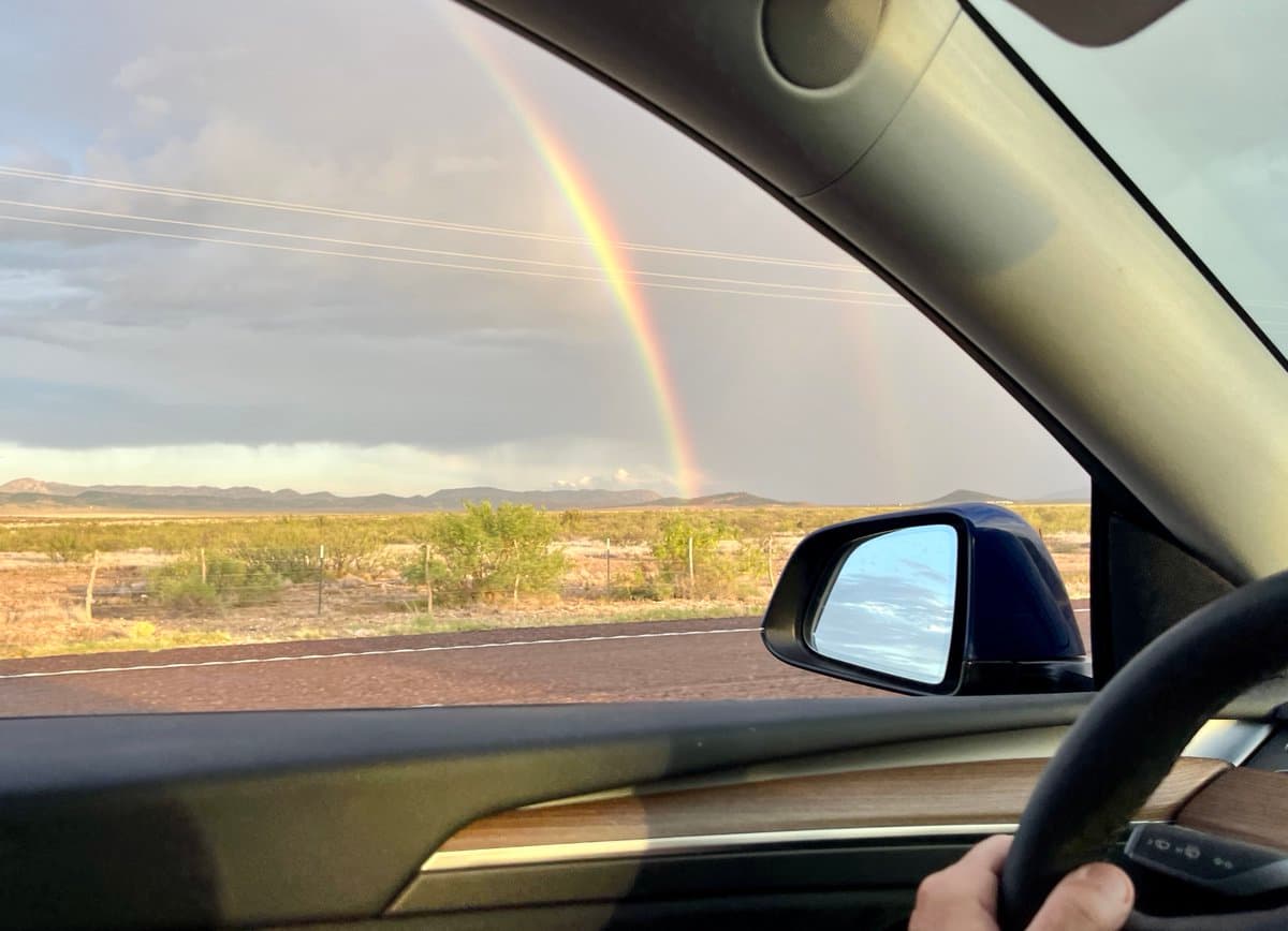 A view from inside a car looking out the passenger window at a desert landscape with a rainbow arching across the sky. The car's side mirror, door panel, and a hand on the steering wheel are visible in the foreground.
