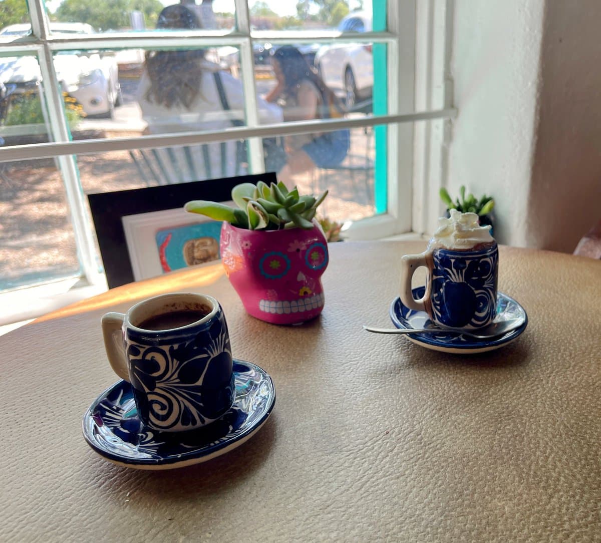 A table inside a cafe holds two blue-and-white patterned ceramic cups of coffee on matching saucers, with a small pink sugar-skull planter holding a succulent in the center. Through the window behind the table, a street scene with parked cars and pedestrians is visible.