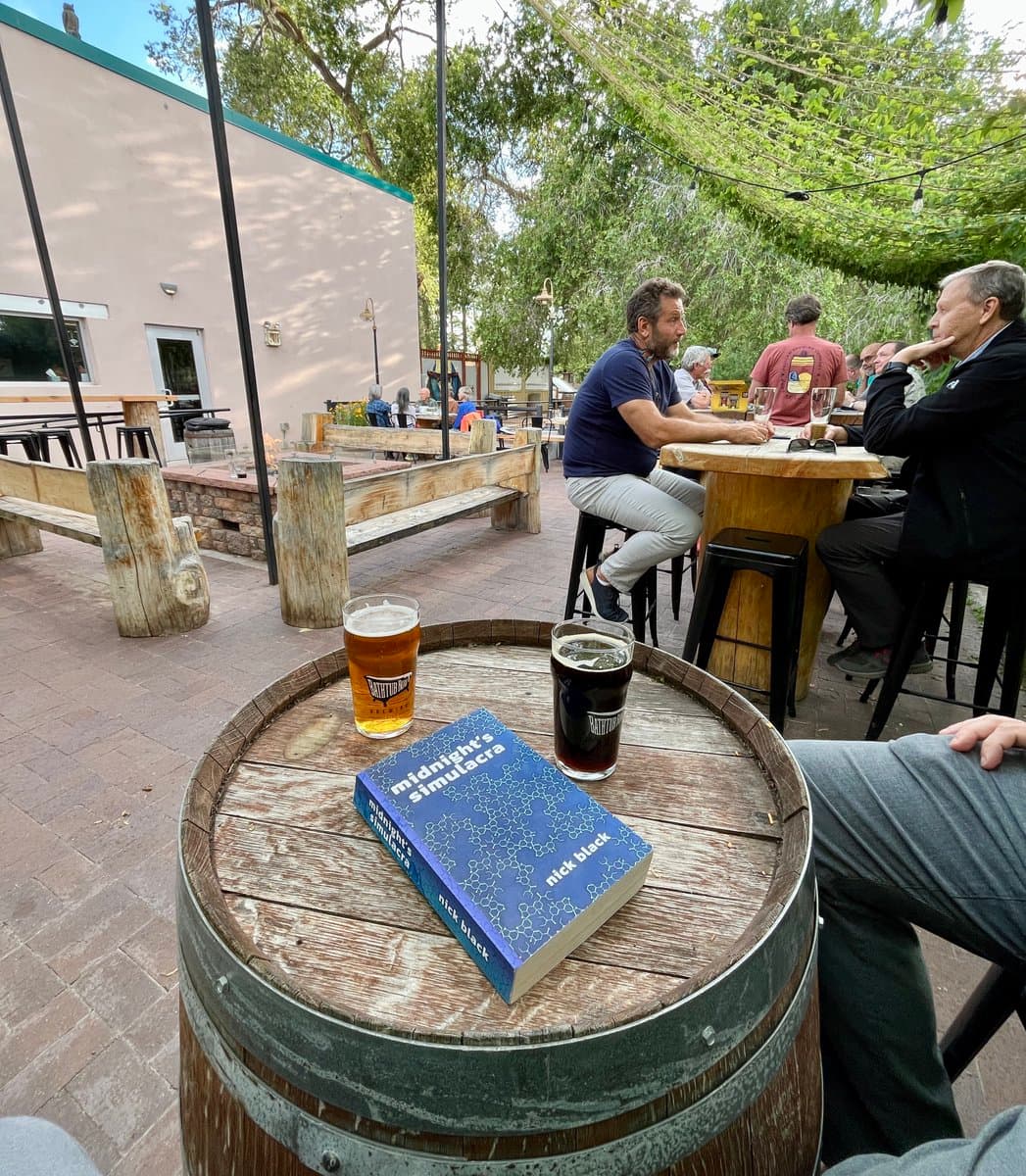 A photograph taken at an outdoor patio, showing a copy of the book "Midnight Simulacra" by Nick Black resting on a wooden barrel table next to two glasses of beer. In the background, several people are seated at high-top tables under a canopy of trees.