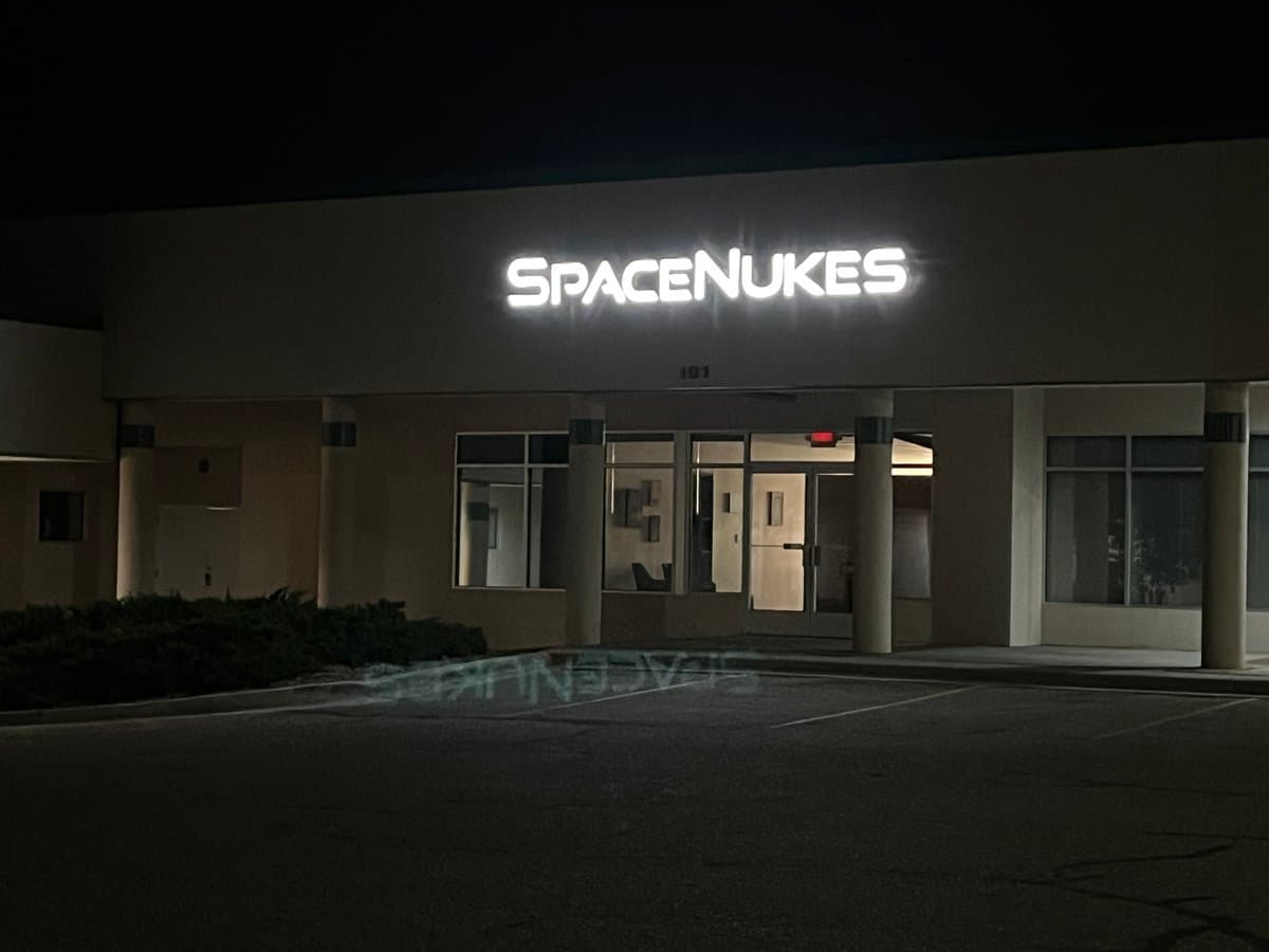 A nighttime photograph of a commercial building with a brightly illuminated sign reading "SPACENUKES" above the entrance. The building features a modern design with large windows and a covered entryway, and the sign's light reflects on the wet pavement in the foreground.