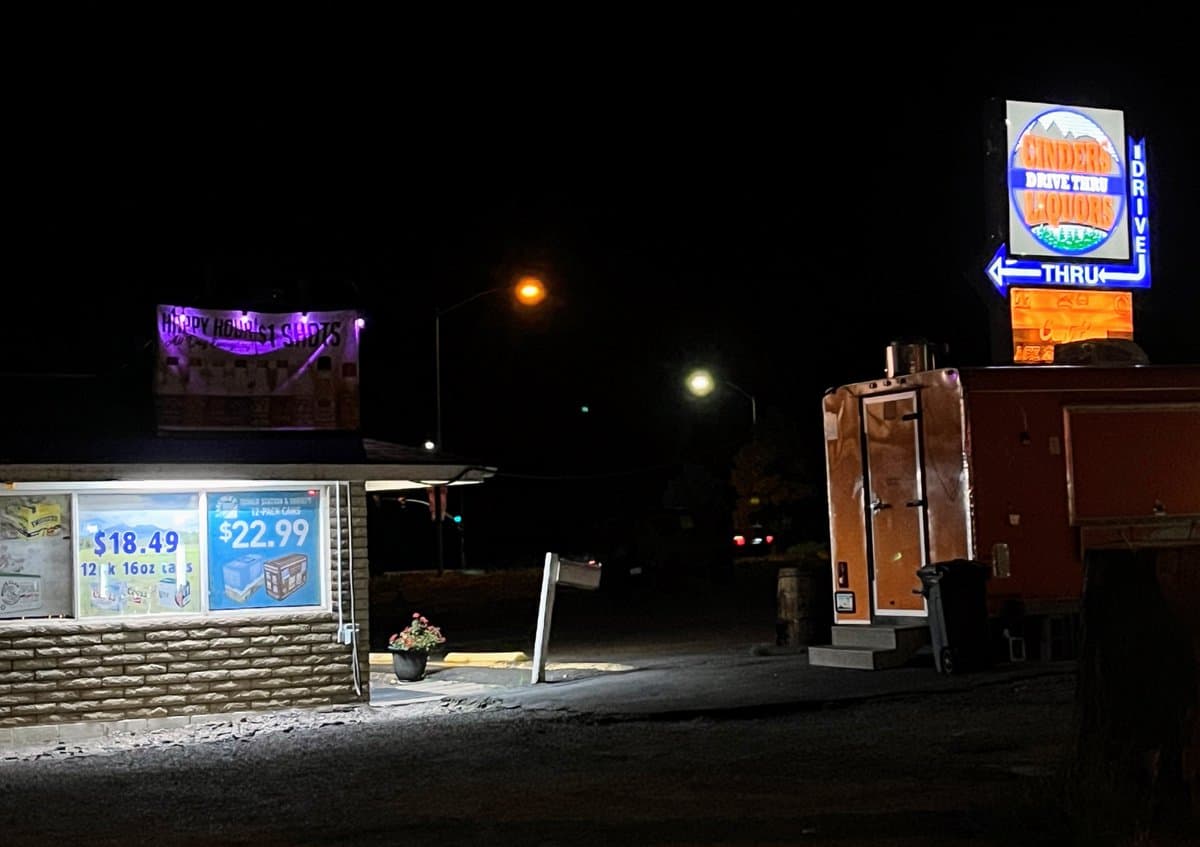 A nighttime photograph of a drive-through liquor store featuring illuminated signage and window advertisements. The scene includes a large neon sign for the business and a banner advertising a happy hour special.
