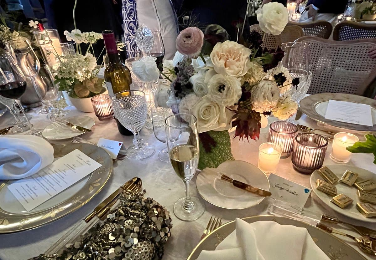A close-up photograph of an elegantly set wedding reception table featuring a centerpiece of white and cream roses, crystal glassware, gold-rimmed plates, and lit candles. The table is decorated with sequined accents and place cards, creating a luxurious and romantic atmosphere.