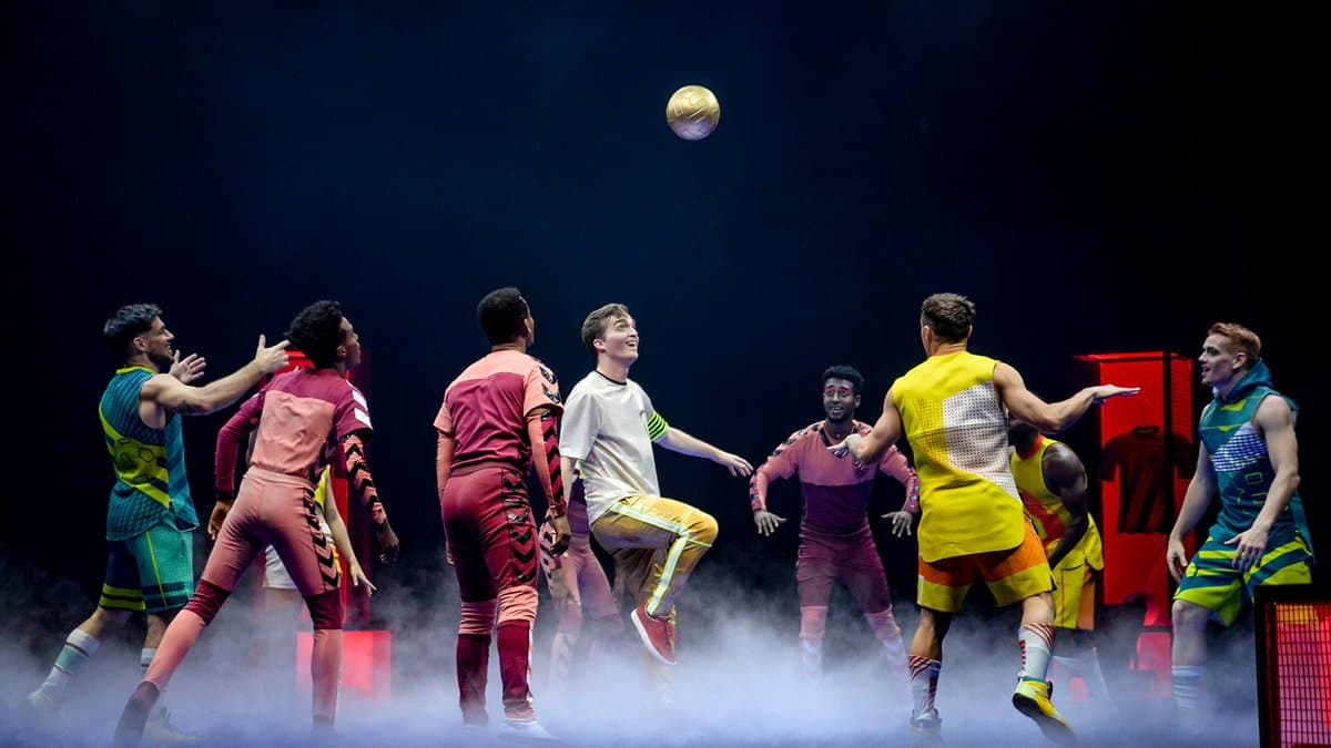 A group of performers in colorful soccer uniforms are on a stage with low-lying fog, watching a golden ball float in the air above them.