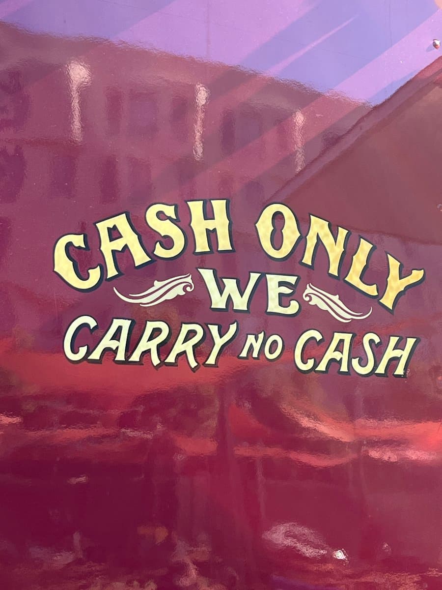 A photograph of a sign on a glass window that contains a contradictory message. The sign features cream-colored text on a dark background with reflections of a building visible on the glass.
