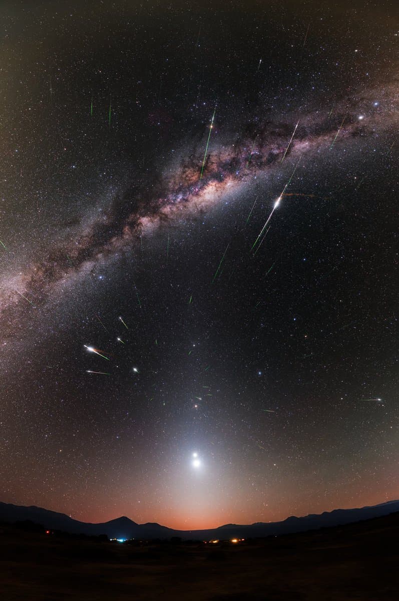 A wide-angle night sky photograph showing the Milky Way galaxy arching across the frame, with multiple bright meteor trails and a bright celestial object near the horizon above a dark mountain silhouette. The image captures a meteor shower event with visible stars and a colorful band of galactic dust and gas. The foreground features a dark landscape with faint lights on the horizon.
