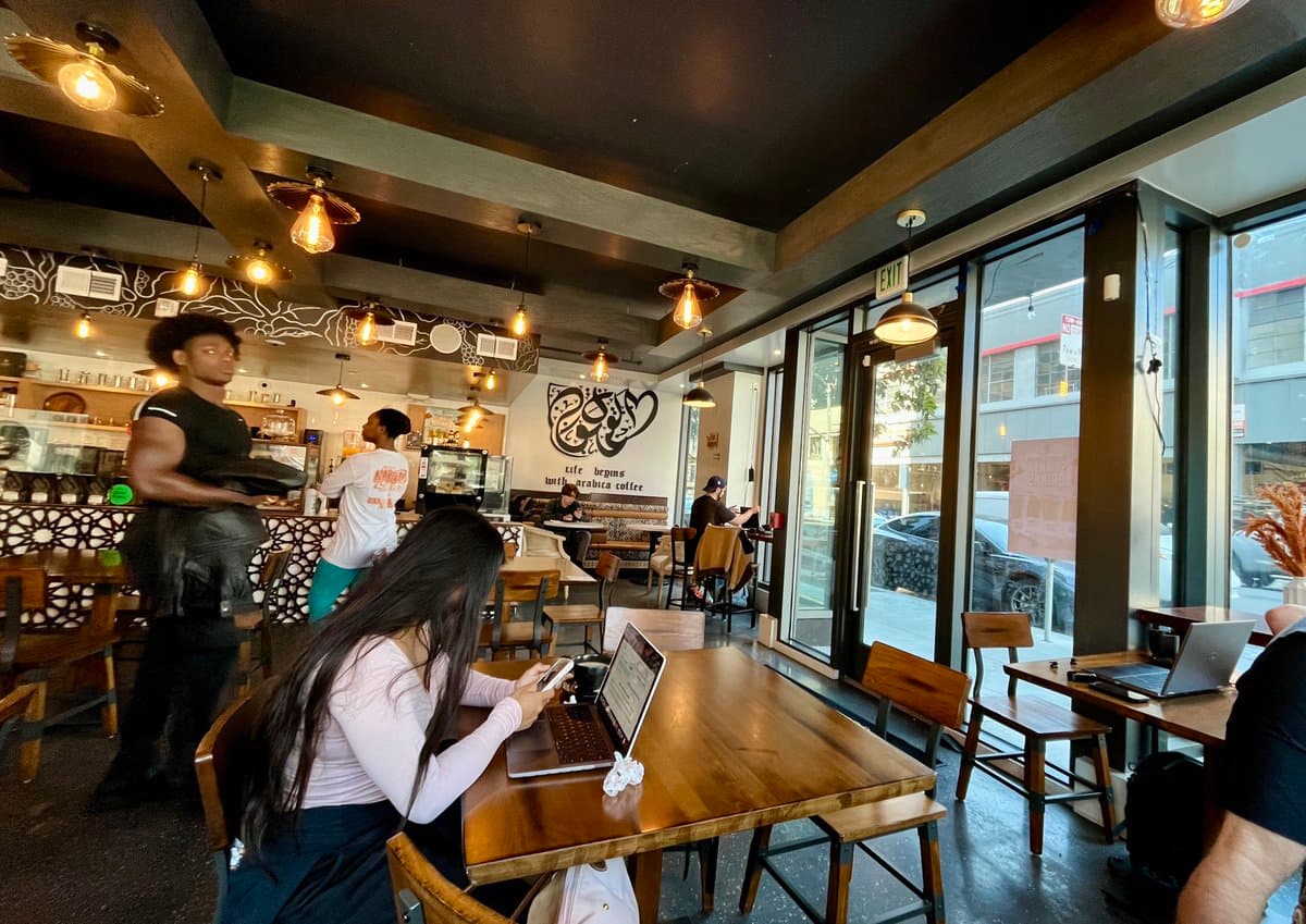 A wide interior shot of a busy coffee shop with wooden tables and hanging light bulbs. A woman in the foreground uses a laptop and phone, while a barista works at the counter in the background.
