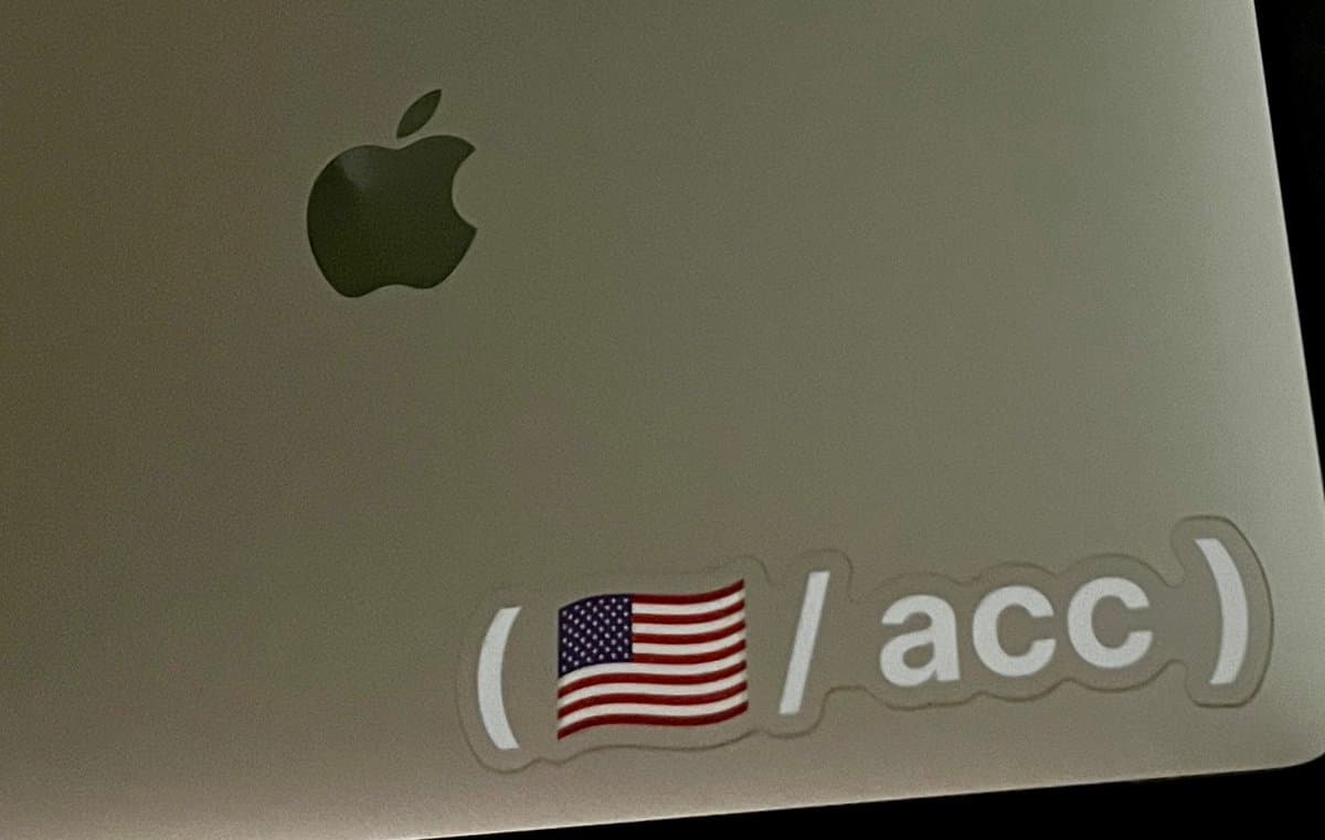 A close-up photograph of the back of a silver Apple laptop, featuring the Apple logo and a sticker with a US flag and the text '/acc'.