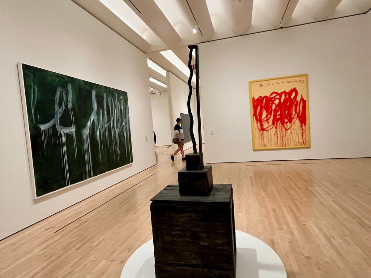 A wide-angle photograph of a contemporary art gallery featuring a large green painting with white drips on the left, a red abstract painting on the right, and a tall, dark sculptural installation in the center. A person is walking in the background between the artworks.