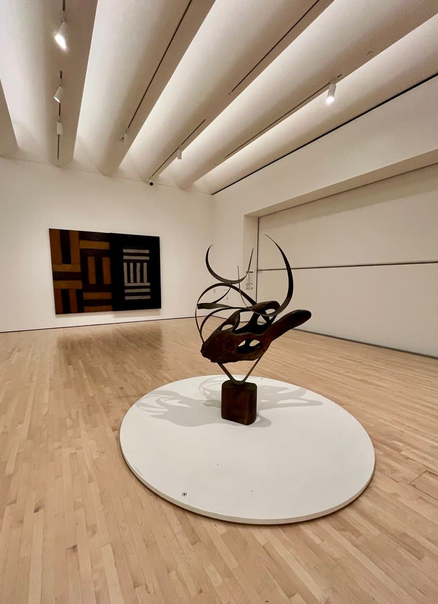 A photograph of a modern art gallery featuring a dark, abstract metal sculpture on a circular white platform in the foreground, with a large geometric painting on the far wall. The room has light wood flooring and a white ceiling with track lighting.