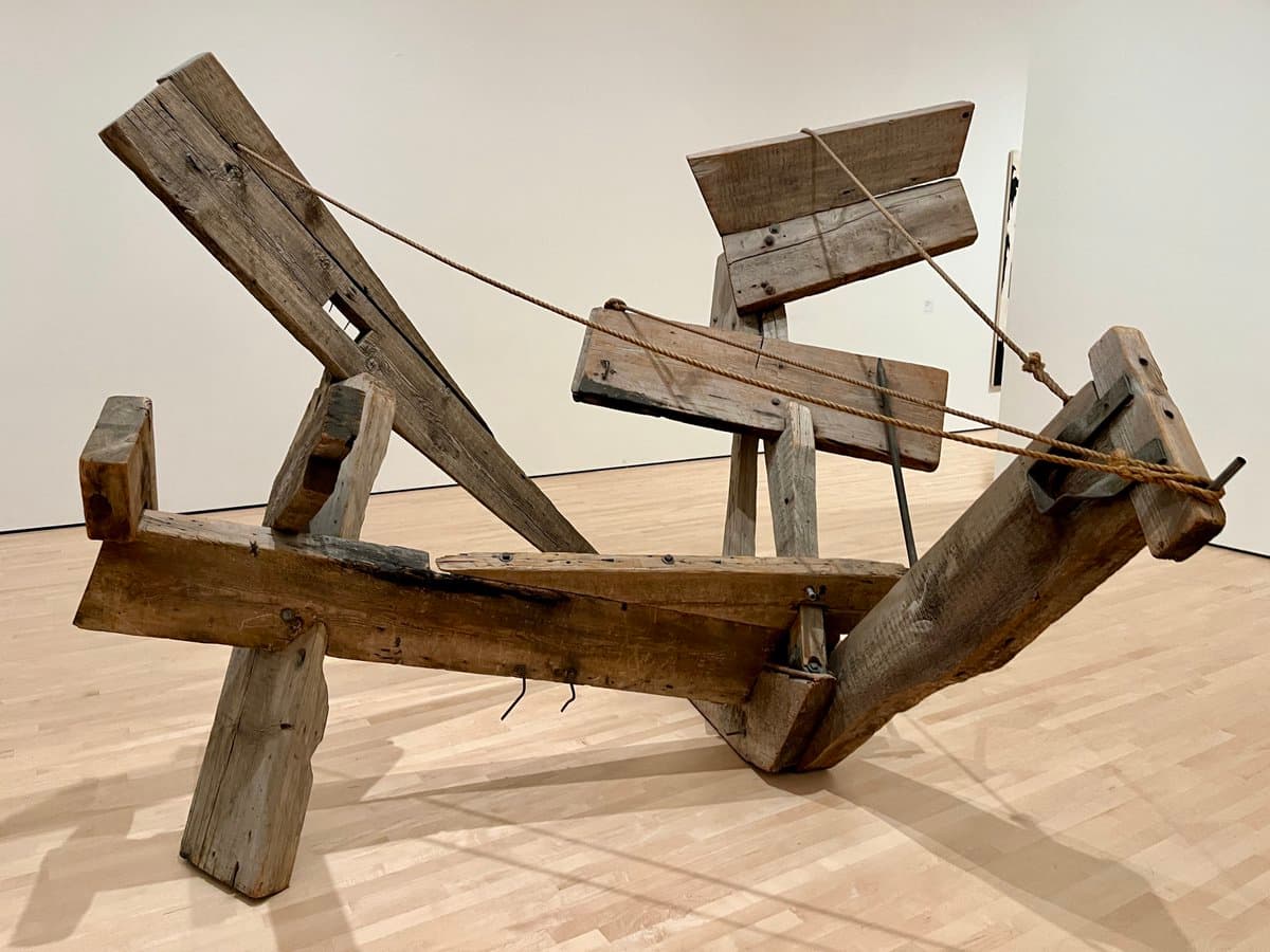 A large, abstract sculpture made of weathered wooden planks and beams, connected by ropes, is displayed on a light wooden floor in a white-walled gallery space. The structure features angled and horizontal elements, creating a dynamic, three-dimensional form.