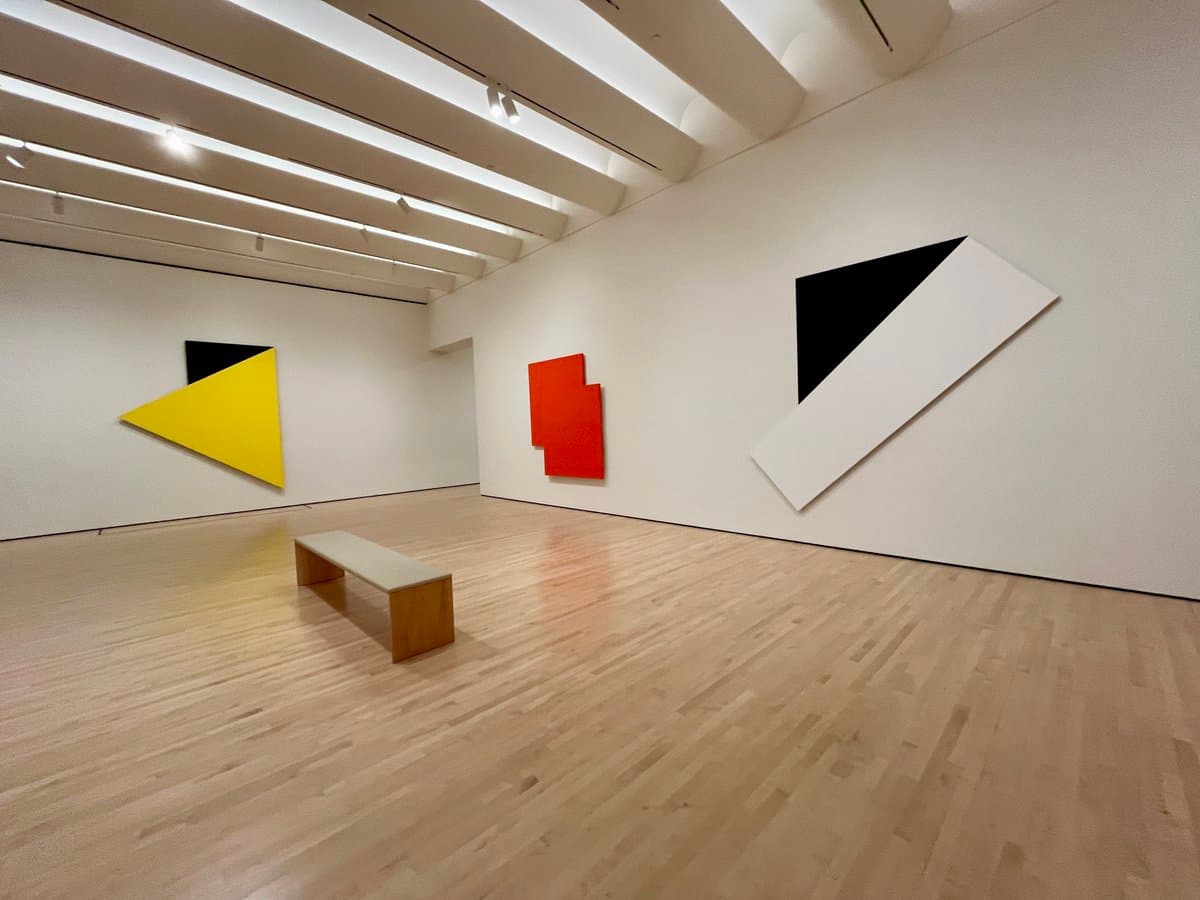 A wide-angle photograph of a minimalist art gallery with white walls and a light wood floor, featuring three large, geometric abstract paintings in black, yellow, orange, and white. A simple wooden bench sits in the center of the room.