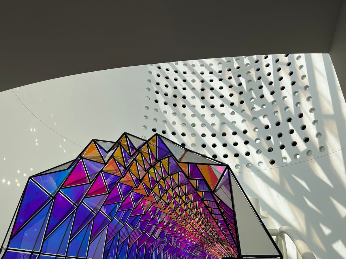 A low-angle photograph of a large, colorful geometric stained-glass sculpture featuring a spectrum of blue, purple, and orange triangular panels. The sculpture is set against a curved white ceiling with a pattern of circular perforations that cast dappled shadows on the wall behind it.