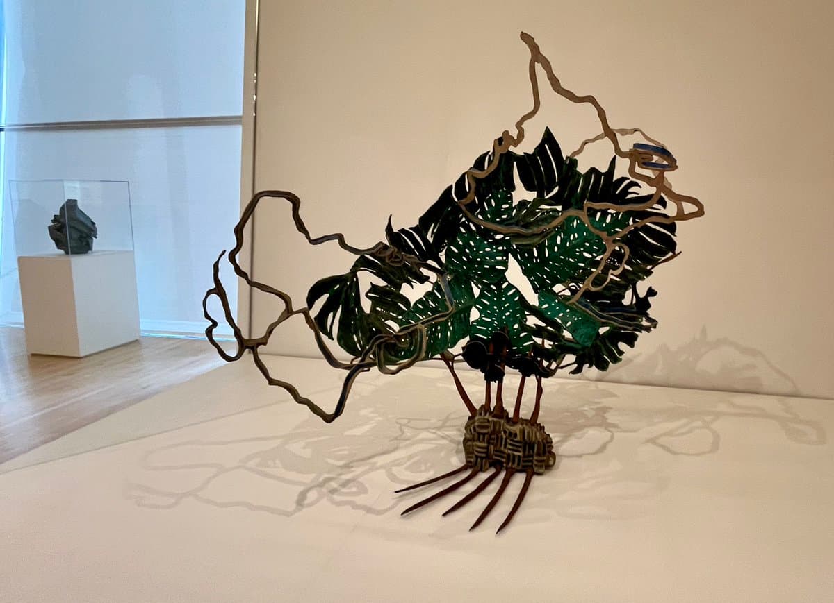 A photograph of a mixed-media sculpture in a gallery setting, featuring a woven base, thin brown wire branches, and green monstera leaves forming a bird-like shape.